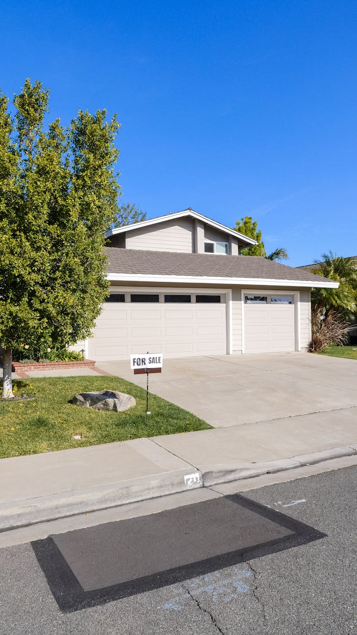 Suburban house with garage and For Sale sign in front yard against clear blue sky.
